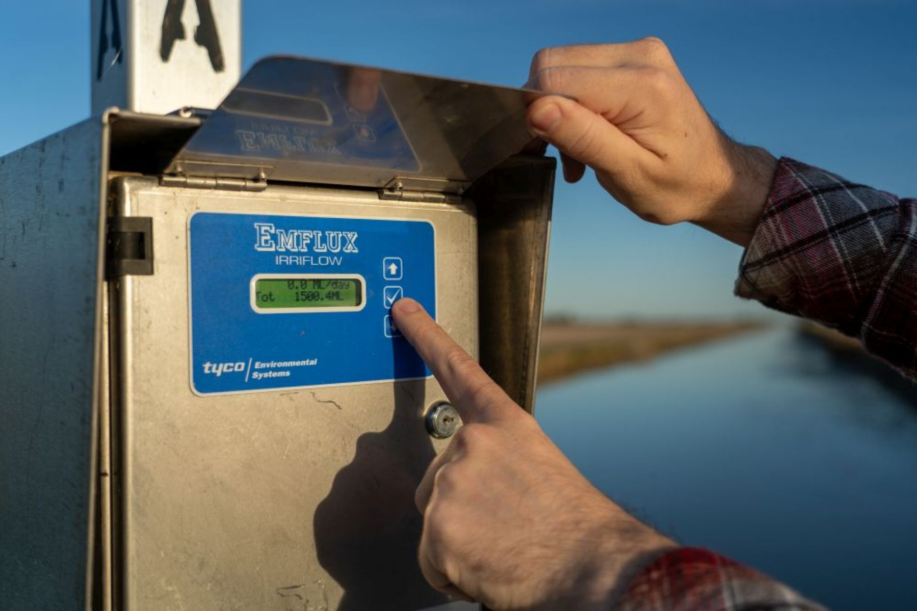 Close-up of a finger pointing at the screen of a metal water meter with a river in the background.